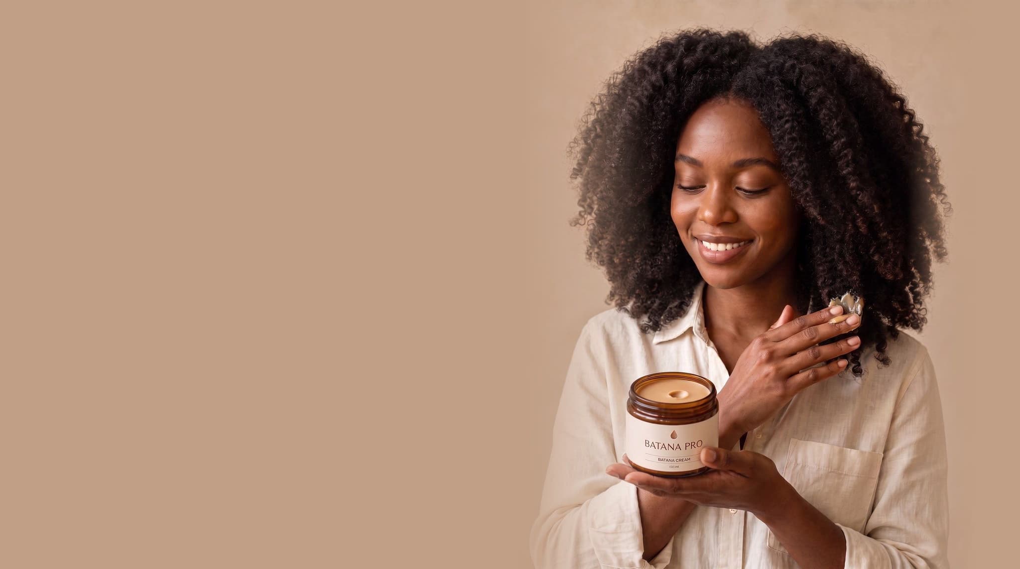 A woman with full, glossy coily hair smiling down at an open jar of Batana Cream, a little on her fingertips