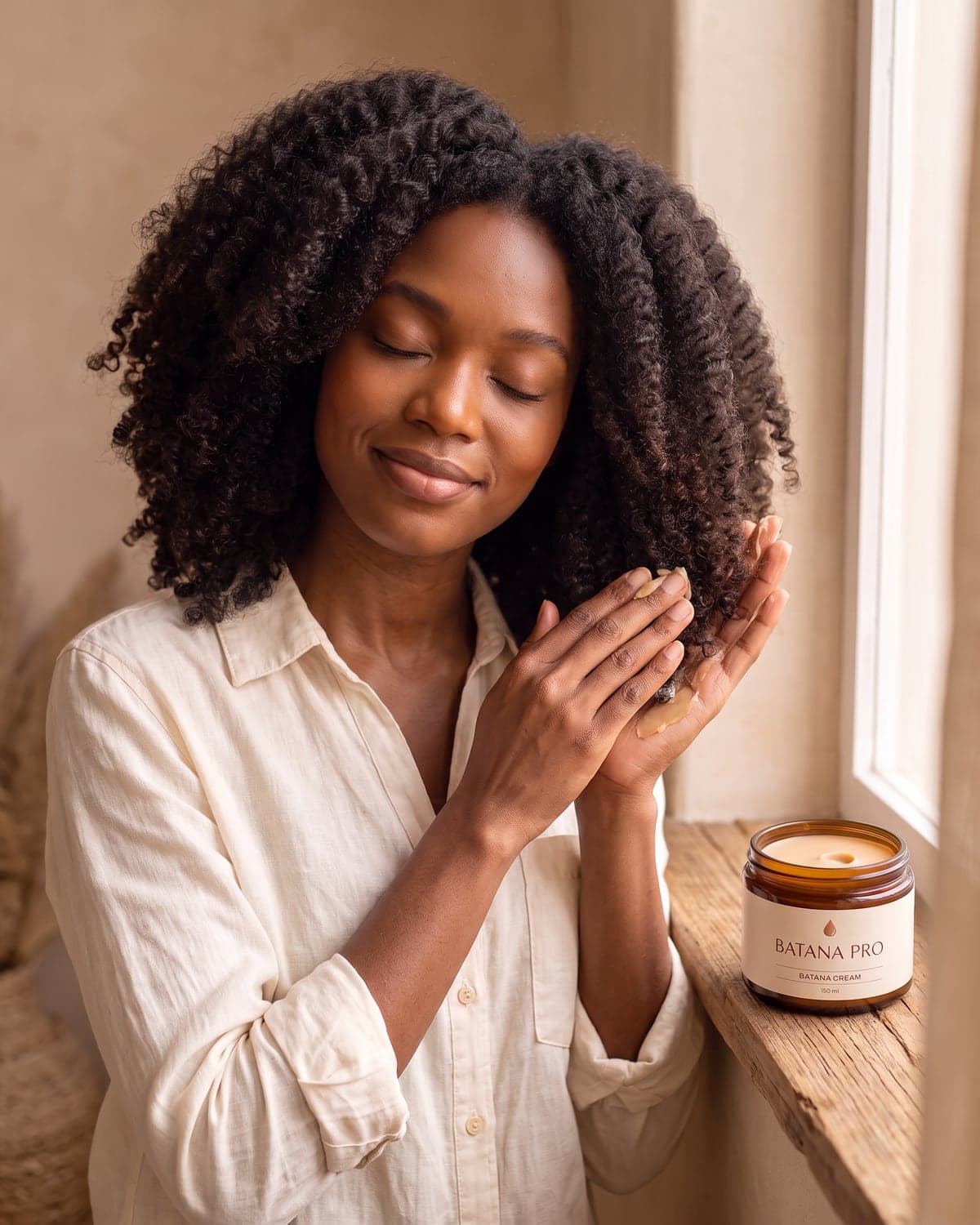 A woman pressing Batana Cream into the ends of her coily hair, eyes closed