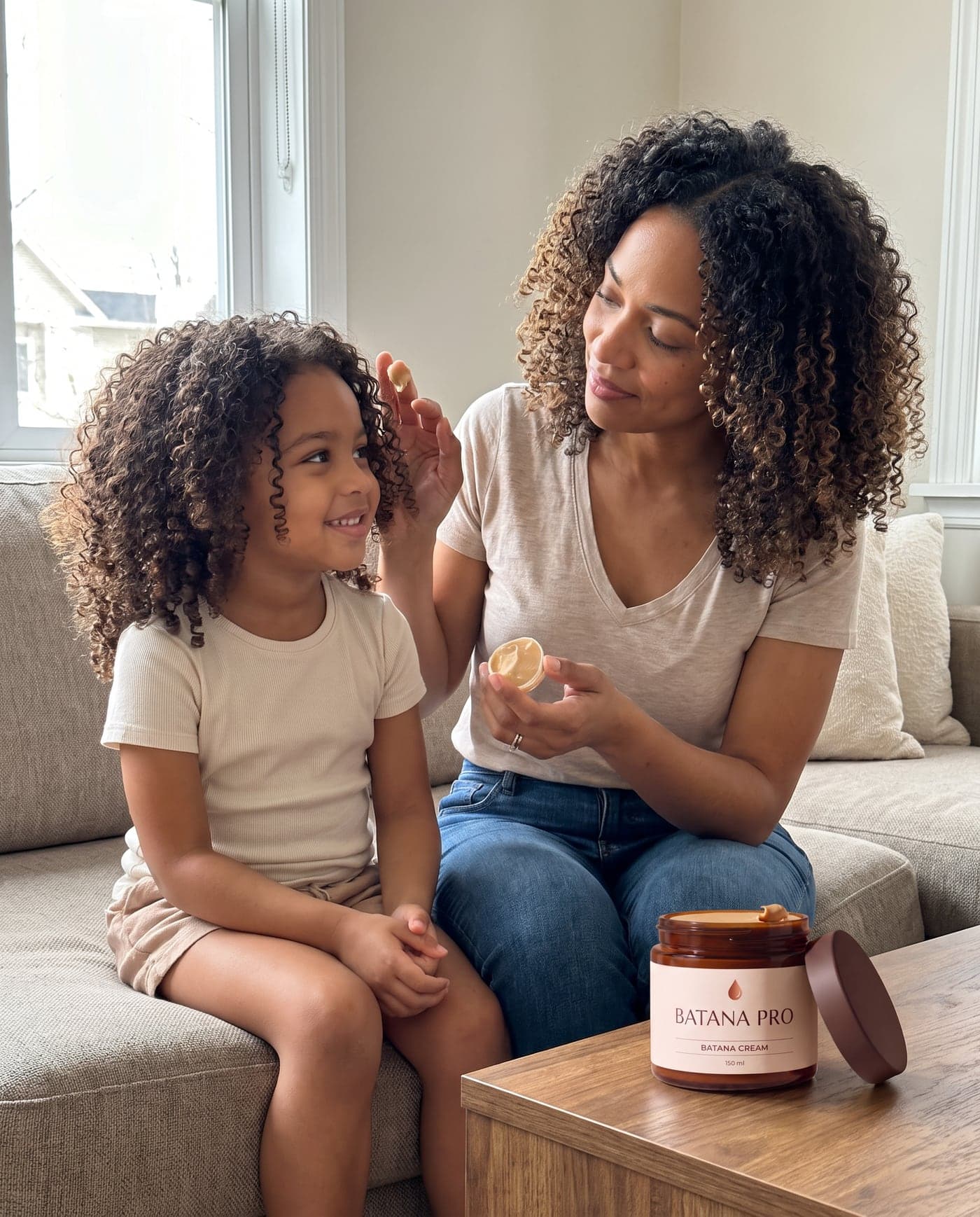 Eine Mutter trägt auf dem Sofa Batana-Creme auf die Locken ihrer Tochter auf